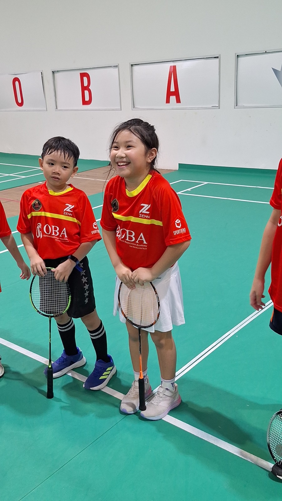 Two children in OBA kit standing together on court holding their rackets